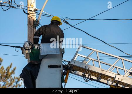 Una vista posteriore di un ingegnere elettrico seduto su una piattaforma fissata con la scala, lavorando con i cavi e riparando qualche guasto tecnico Foto Stock