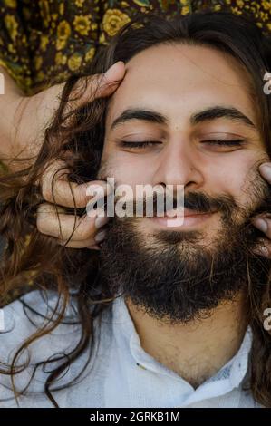 ritratto di un giovane uomo portico e dai capelli lunghi con le mani di una donna carezzandogli la testa e la barba Foto Stock