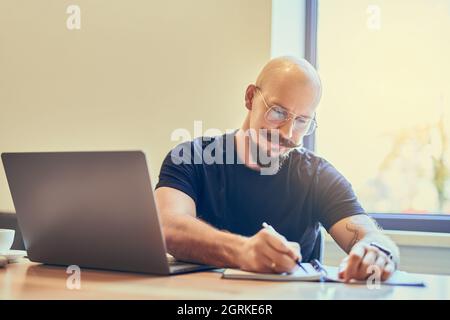 Concentrato uomo uomo calvo adulto che usa il laptop mentre scrive le note nel notebook sul posto di lavoro. Foto Stock