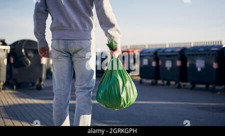 Uomo che cammina con rifiuti. Vista posteriore della persona che trasporta il sacchetto di plastica contro i bidoni della spazzatura sulla strada. Foto Stock