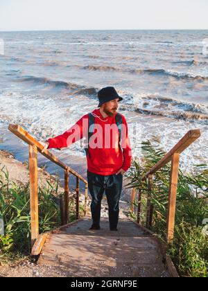 Uomo bearded millennial in rosso Hoody con zaino autunno mare sfondo secco canne autentico maschio turistico lifestyle foto Foto Stock