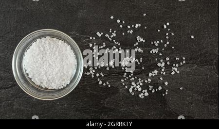 Piccola ciotola di vetro con sale di roccia bianco su un tavolo simile a marmo nero - alcuni cristalli sparsi vicino, vista dall'alto Foto Stock