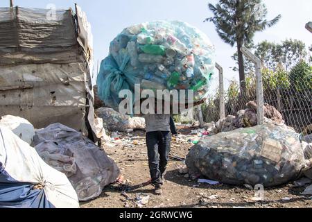 Nakuru, Kenya. 01 ottobre 2021. Un uomo porta un carico pesante di rifiuti di bottiglie di plastica sulle sue spalle a un impianto di riciclaggio vicino al sito di scarico di Gioto a Nakuru. Il problema crescente dei rifiuti di plastica che finiscono nell'ambiente sta diventando una preoccupazione e gli ambientalisti stanno chiedendo maggiori investimenti nelle infrastrutture per riciclare la plastica promuovere l'economia circolare e ridurre l'inquinamento della plastica. Essi chiedono inoltre al governo di introdurre un sistema obbligatorio di deposito e rimborso delle bottiglie (DRS) che darà valore alle bottiglie di plastica per bevande comunemente note come animali da compagnia (polietilentereftalato). De Foto Stock