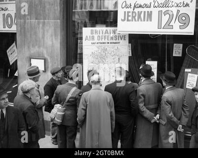 Un aviatore britannico è tra un gruppo di civili affollato intorno alla finestra di un negozio a Holborn, Londra, per guardare una mappa visualizzata che è intitolato 'British Air Offensive' e mostra oltre 700 incursioni da parte della Royal Air Force sulla Germania. Foto Stock