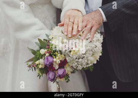 Primo piano delle mani di una sposa e di uno sposo con fedi nuziali, tenendo un bouquet e firmando il certificato di matrimonio ufficiale durante la cerimonia. Foto Stock