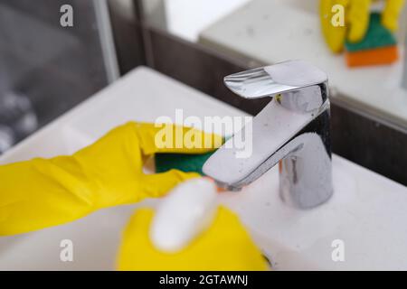 Persona in guanti gialli lava lavandino con panno e spruzza schiuma detergente sul rubinetto Foto Stock