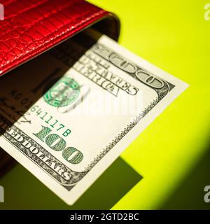 Bolletta del centinaio di dollari che attacca da una borsetta rossa sopra il tavolo verde soleggiato con le ombre forti.