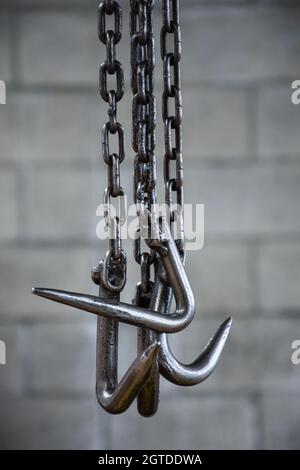 Colpo verticale di ganci metallici appesi alle catene su uno sfondo grigio della parete di mattoni. Foto Stock