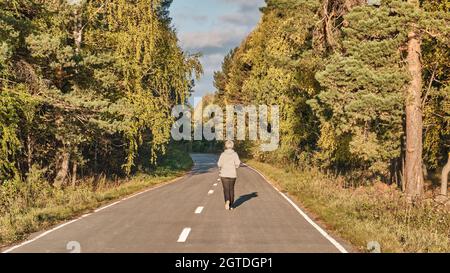 Donna attiva senior con capelli grigi che corrono lungo la strada lastricata foresta il giorno d'autunno soleggiato. Foto Stock