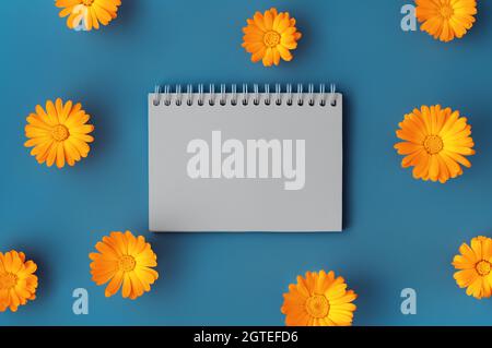 Sfondo bellissimo con blocco note a spirale vuoto aperto incorniciato con fiori arancione marigold motivo su sfondo blu. Sfondo per il tuo progetto. Stile a disposizione piatta. Spazio di copia. Foto Stock