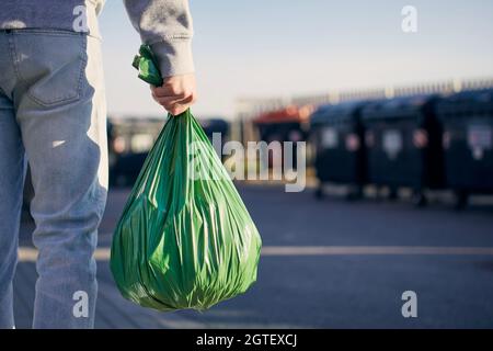 Uomo che cammina con rifiuti. Vista posteriore della persona che trasporta il sacchetto di plastica contro i bidoni della spazzatura sulla strada. Foto Stock