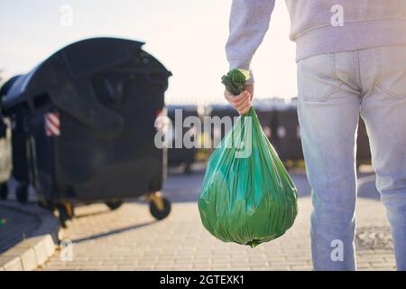 Uomo che cammina con rifiuti. Vista posteriore della persona che trasporta il sacchetto di plastica contro i bidoni della spazzatura sulla strada. Foto Stock