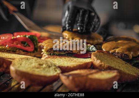 Processo di cottura degli cheeseburgers sul grill. Chef in guanti neri che cucinano un hamburger Foto Stock