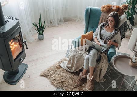 Lavoro a distanza. Giovane donna che usa un computer portatile in una comoda poltrona e plaid seduta accanto al camino con un gatto domestico, bere il tè a casa Foto Stock