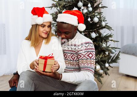 Momenti accoglienti e gioiosi nelle vacanze invernali. Una coppia multinazionale apre un regalo di Natale. Un giovane uomo nero amorevole e una donna bianca sono seduti accanto Foto Stock