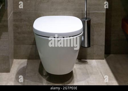 WC dal design interno moderno, WC bianco sospeso con coperchio chiuso e spazzola. Accessori da bagno di lusso su pareti piastrellate in marmo e pavimento Foto Stock