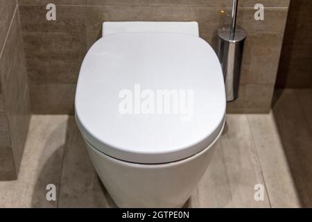 WC dal design interno moderno, WC bianco sospeso con coperchio chiuso e spazzola. Accessori da bagno di lusso su pareti piastrellate in marmo e pavimento Foto Stock