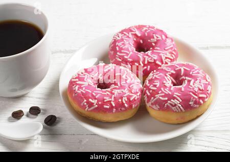 Ciambelle rosa con spruzzi in piatto e tazza di caffè su tavola bianca in legno Foto Stock