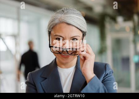Felice donna d'affari matura con capelli bianchi in occhiali sorridenti alla macchina fotografica in ufficio Foto Stock