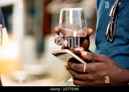 mano di un giovane africano che tiene un bicchiere di vino rosso e un telefono cellulare Foto Stock