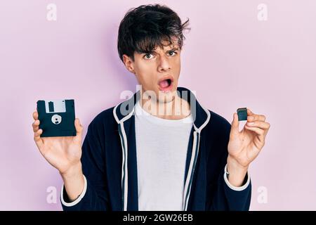 Bell'hipster giovane uomo che tiene il floppy disk e la scheda sdxc in faccia d'urto, guardando scettico e sarcastico, sorpreso con bocca aperta Foto Stock