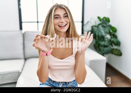 Giovane donna bionda che tiene invisibile allineatore ortodontico sorridente allegro presentando e indicando con palmo di mano guardando la macchina fotografica. Foto Stock