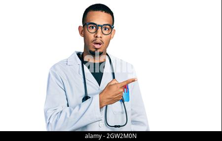 Giovane uomo africano americano che indossa uniforme medico e stetoscopio sorpreso puntando con il dito di lato, bocca aperta espressione stupita. Foto Stock