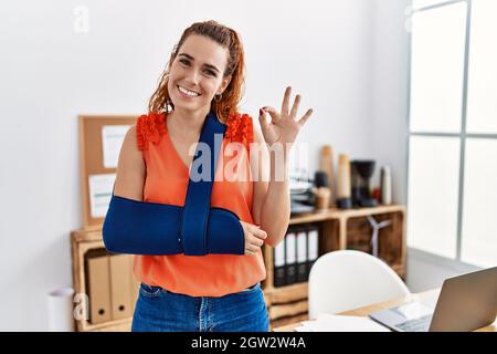 Giovane donna rossa che indossa braccio su imbracatura in ufficio facendo ok segno con le dita, sorridendo amichevole gesturing eccellente simbolo Foto Stock