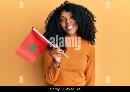 Donna afroamericana con capelli afroamericani che reggono la bandiera del marocco guardando positivo e felice in piedi e sorridendo con un sorriso sicuro mostrando i denti Foto Stock