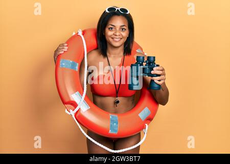 Giovane ragazza afroamericana che tiene il galleggiante del bagnino usando binocoli sorridendo con un sorriso felice e freddo sul viso. Mostrando i denti. Foto Stock