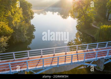 Ponte di legno con corrimano bianchi sul fiume. I raggi del sole brillano attraverso la nebbia mattutina. Foto Stock