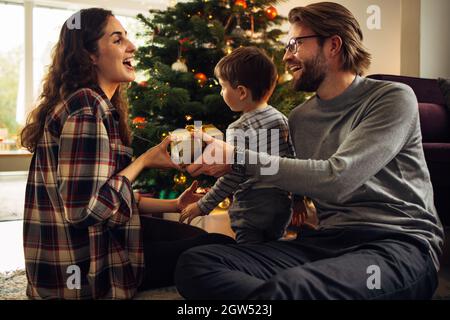 Donna che riceve un regalo per natale e Capodanno da suo marito. Felice giovane famiglia seduta da albero di Natale la vigilia di natale. Foto Stock