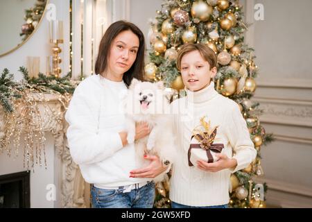 Felice famiglia caucasica madre e figlio seduto a casa vicino albero di natale con il loro cane piccolo. Ritratto di famiglia a Capodanno e a natale. Si danno a vicenda doni, volti felici. In un maglione bianco Foto Stock