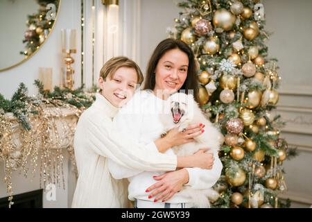 Bella mamma e figlio sorridente che abbraccia un cane bianco felice a casa per le vacanze di natale. Famiglia felice sorridendo e festeggiando il natale con il loro cane divertente. Vacanze di Natale Foto Stock