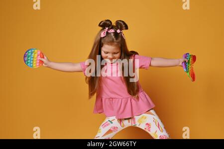 Ragazza di capretto frolic in vestiti colorati tiene due giocattoli sensoriali di colore dell'arcobaleno - schioccalo e guarda verso il basso. Giocare e ballare Foto Stock