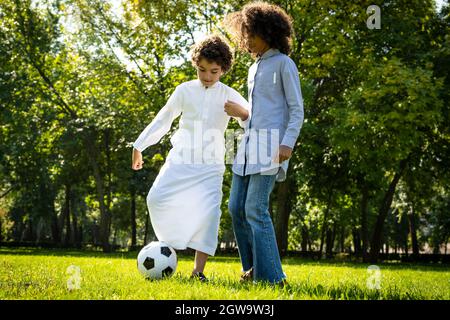 immagine cinematografica di una famiglia degli emirates che trascorre del tempo al parco. Fratello e sorella che giocano a calcio in erba Foto Stock