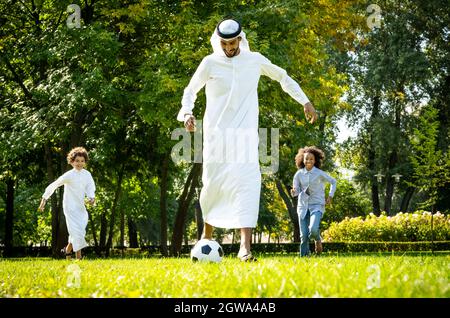 immagine cinematografica di una famiglia degli emirates che trascorre del tempo al parco. Fratello e sorella che giocano a calcio in erba Foto Stock