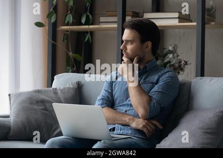 Uomo premuroso seduto sul divano con un computer portatile, guardando a distanza Foto Stock