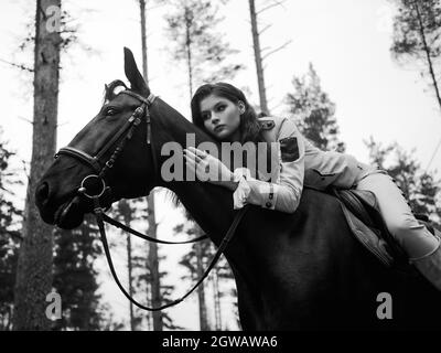 Un giovane e bellissimo pilota di brunette in un elegante abito retrò seduto a cavalcare un cavallo nero appoggiato sul collo. Tinta bianca e nera Foto Stock