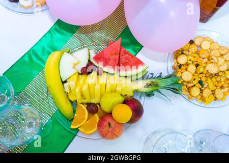 Piatto con ananas a forma di pesce, melone, cocomero, uva, arance, pera, mela e banana su un tavolo, accanto ad esso un piatto con noci e som Foto Stock