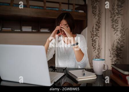La studentessa asiatica, dubbia e premurosa, che guarda via pensando di risolvere il problema, sente la mancanza di nuove idee creative sul lavoro, pensivo puzzled o annoiato, si siede alla scrivania con un computer portatile Foto Stock