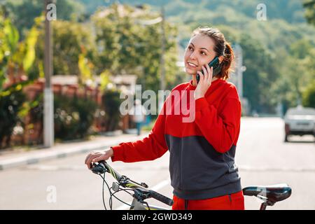 Comunicazione. Un ritratto di giovane bella donna sorridente sta facendo una telefonata e spingendo una bicicletta. Strada soleggiata sullo sfondo. All'aperto. Modalità Foto Stock
