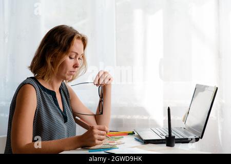 Una giovane donna seduta alla scrivania, che controlla il lavoro dei bambini. Sul tavolo sono presenti un computer portatile e disegni. Il concetto di autoisolamento e di freelance. Foto Stock