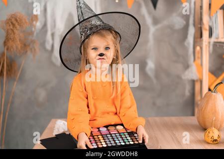 Halloween trucco bambina nella stanza sul tavolo con una tavolozza di ombre nelle sue mani si sta preparando per la vacanza. Un bambino in un cappello strega. Foto Stock