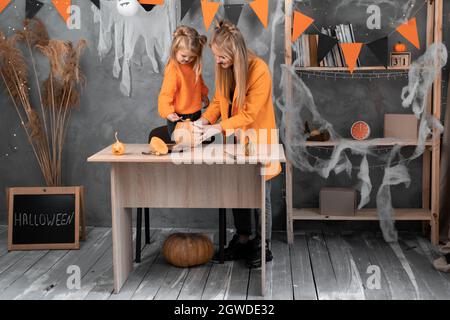 Mamma e figlia della famiglia preparano la zucca per Halloween a casa in una camera arredata. Le ragazze bionda stanno ordinando una zucca con un cucchiaio. famiglia di halloween Foto Stock