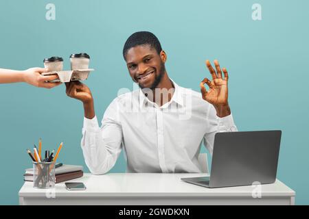 Uomo d'affari nero che riceve caffè da asporto o tè nel portabicchiere e che mostra il cartello ok, seduto alla scrivania con il computer portatile isolato su sfondo blu. Cibo de Foto Stock