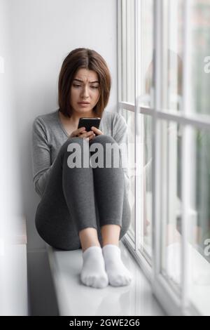 Triste sconvolto caucasico millennial femmina seduta sul davanzale a casa e leggere messaggio al telefono con cattive notizie, sofferente di depressione. Reazione a. Foto Stock