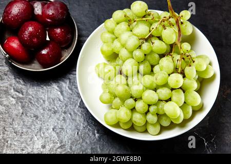 Ramo di uva verde matura sul piatto con gocce d'acqua e prugne in ciotola. Frutta succosa su sfondo di legno, primo piano. Cibo sano sul tavolo da cucina scuro Foto Stock