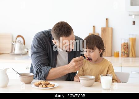 Nutrizione dei bambini. Amando papà che nutriva il figlio con porridge, seduto in cucina al mattino, uomo che si prende cura del suo bambino, spazio libero Foto Stock
