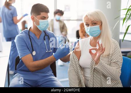 Donna matura felice in maschera di faccia che mostra il gesto giusto segno mano mentre si riceve il vaccino nel centro di salute. Vaccinazione, immunizzazione, inoculatio Foto Stock